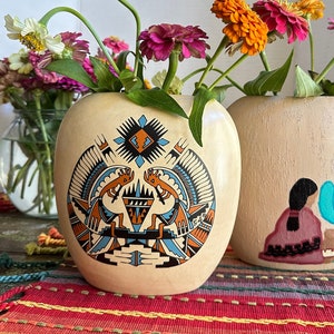 May include: Two beige, gourd-shaped vases with colorful floral arrangements. One vase features a Native American-inspired design in blue, orange, and black. The other vase has a painted figure and a turquoise cactus. The vases sit on a colorful woven mat.