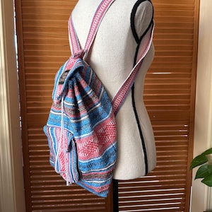 May include: A woven backpack featuring a vibrant striped pattern in shades of blue, pink, and white. The bag includes adjustable shoulder straps, a drawstring closure, and a front pocket with a black rectangular label displaying white text.