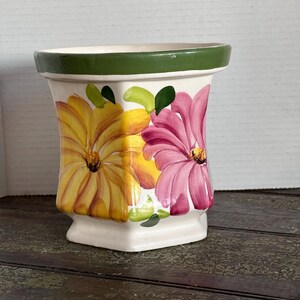 May include: A ceramic planter with a cream-colored base and a wide green rim. The exterior features hand-painted floral motifs in shades of yellow and pink, accented with green leaves. The vessel has a flared top and a square-shaped base.