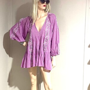 May include: A purple mini dress with white embroidered details. The dress has a V-neck, long sleeves, and a tiered skirt. The dress is made of a striped fabric and is displayed on a mannequin. The mannequin is wearing sunglasses.