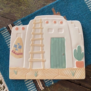 May include: A white ceramic Southwestern-style decorative piece featuring a pueblo design with a ladder, door, and cactus. The piece has a turquoise door and accents, with a tan base and a small hanging textile. It rests on a blue and white woven textile.