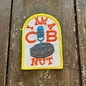 May include: Embroidered patch with a yellow border and the words "I AM A COB NUT" in orange and white. The patch features a blue microphone design above a gray, hexagonal shape. The patch is on a weathered wooden surface.