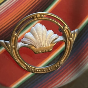 May include: A vintage-style brooch with a gold-colored metal frame. The brooch features a white, shell-shaped centerpiece and a decorative circular design. The background is a colorful striped pattern of red, orange, green, and blue.