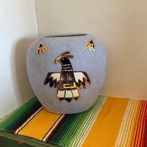 May include: A light blue ceramic vase with a stylized bird design in black, white, and gold. The vase has a rounded shape and features additional decorative elements. The vase is displayed on a colorful striped textile.