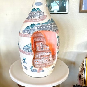 May include: A cream-colored ceramic table lamp with a textured surface featuring painted desert landscapes and a cutout section revealing a three-dimensional terracotta-colored pueblo dwelling illuminated from within by a warm light.