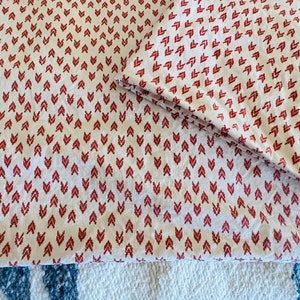 May include: White fabric with a repeating pattern of red arrow-like shapes. The fabric is folded, showing the pattern on the top and the folded edge. The fabric is likely a sheet or bedding material.