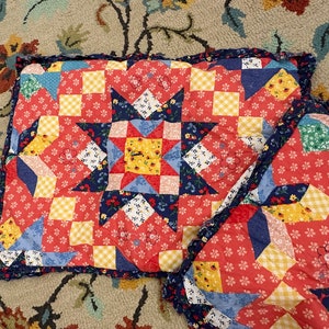May include: Handmade patchwork quilt with a star design. The quilt features a red background with blue, yellow, and white floral and geometric patterns. The edges are finished with a ruffled blue border. A cozy, decorative home textile.