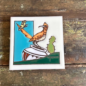 May include: Square ceramic tile featuring a colorful illustration of a roadrunner bird perched on a decorative pot. The design includes a cactus, a butterfly, and a turquoise background. The tile is white with a green border and the artist's signature.