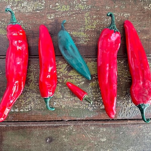 May include: An overhead shot of five ceramic chili peppers on a weathered wooden surface. Four are bright red with green stems, and one is teal. One small red pepper is also included.