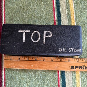 Op de afbeelding: Een zwarte, rechthoekige olie-steen met het woord "TOP" in witte hoofdletters. De woorden "OIL STONE" zijn ook zichtbaar. Een houten liniaal ligt naast de steen, met metingen in centimeters.