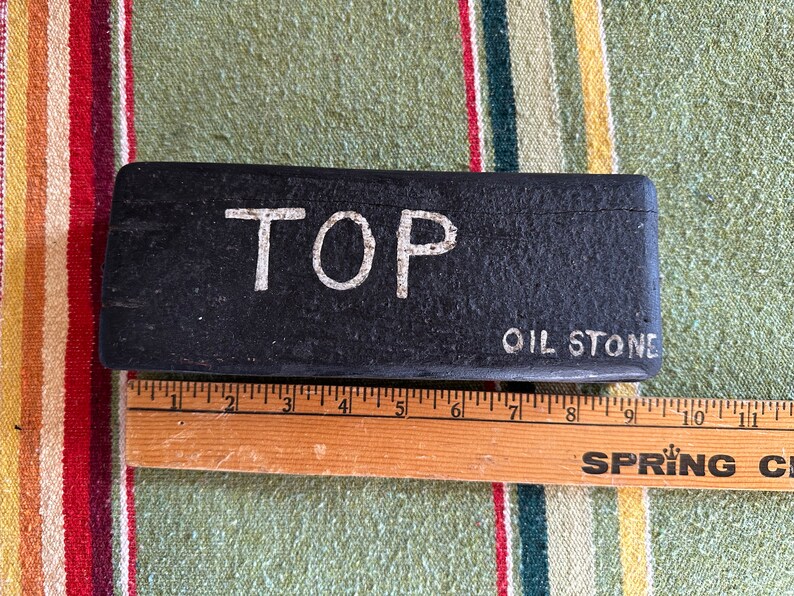 Op de afbeelding: Een zwarte, rechthoekige olie-steen met het woord "TOP" in witte hoofdletters. De woorden "OIL STONE" zijn ook zichtbaar. Een houten liniaal ligt naast de steen, met metingen in centimeters.