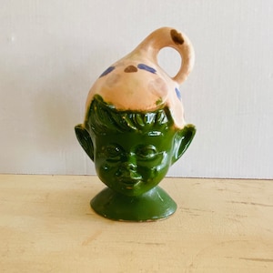 May include: A whimsical ceramic figurine in the shape of a face with a hat. The face is a vibrant green, while the hat is a light tan with blue and brown accents. The figurine has a loop on top and is set on a small base.