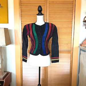 May include: A black long-sleeve jacket featuring vertical stripes in red, green, and blue across the front. The jacket has a round neckline, a front button closure, and horizontal red and yellow stripes on the cuffs of both sleeves.