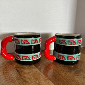 May include: Two ceramic mugs with a black base, adorned with turquoise and red geometric patterns. The handles are shaped like red chili peppers. The mugs are designed with a unique and eye-catching aesthetic.