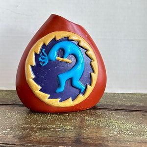 May include: A terracotta-colored vase featuring a circular medallion with a jagged gold border. Inside the circle, a bright blue Kokopelli figure plays a flute against a dark purple background. The vase has a wide, rounded base and a slanted top opening.