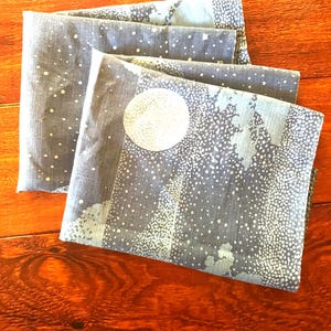 May include: Folded gray fabric with a white dotted pattern and a large white circle design. The fabric has a light blue border and is stacked on a wooden surface. The design evokes a night sky or winter scene.