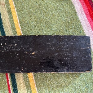 Op de afbeelding: Een rechthoekig, zwart houten object met een verweerde afwerking. Het hout vertoont tekenen van slijtage, met zichtbare nerven en afgebladderde verf. Het object staat tegen een gestreepte, kleurrijke textielachtergrond.