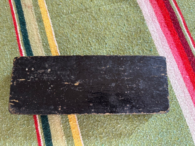 Op de afbeelding: Een rechthoekig, zwart houten object met een verweerde afwerking. Het hout vertoont tekenen van slijtage, met zichtbare nerven en afgebladderde verf. Het object staat tegen een gestreepte, kleurrijke textielachtergrond.
