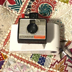 May include: A vintage Polaroid Swinger Land Camera, model 20, with a silver and black body, red shutter button, and a black lens. The camera sits on a white base. The image is set against a colorful, patterned fabric.