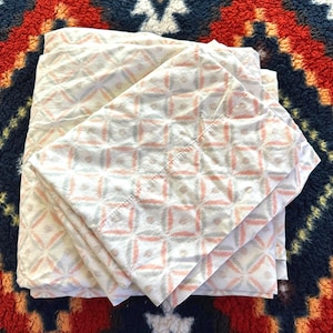 May include: Folded white bed sheets with a repeating geometric pattern in coral, gray, and blue. The sheets are neatly stacked on a patterned rug with red, navy, and white colors.