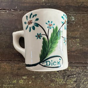 May include: White ceramic mug with a curved handle, decorated with teal flowers and green leaves. The word "Dick" is written in black script on the front of the mug. The mug is set against a rustic wooden surface.