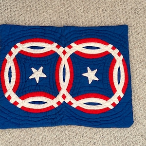 May include: A blue, red, and white quilted pillow with a patriotic design. The pillow features two overlapping circles with red and white stripes, white stars, and a blue background. The quilting creates a textured effect.