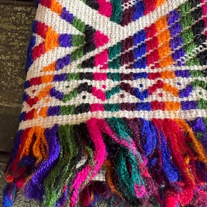 May include: Close-up of a colorful woven textile with a geometric pattern. The design features a cream-colored base with vibrant accents in orange, purple, green, pink, and blue. The bottom edge has a fringe of multicolored yarn. The textile is likely a scarf or shawl.