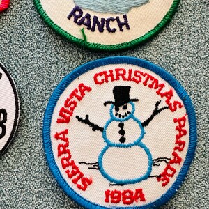 Op de afbeelding: Geborduurde patch met een blauwe rand met een sneeuwpop met een zwarte hoge hoed, met de tekst "Sierra Vista Christmas Parade 1984" in het rood. De sneeuwpop heeft zwarte armen en een kolenachtig knoopontwerp.