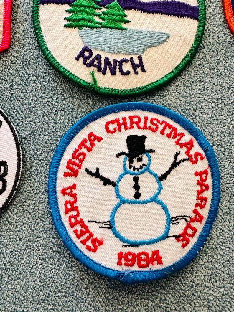 Op de afbeelding: Geborduurde patch met een blauwe rand met een sneeuwpop met een zwarte hoge hoed, met de tekst "Sierra Vista Christmas Parade 1984" in het rood. De sneeuwpop heeft zwarte armen en een kolenachtig knoopontwerp.