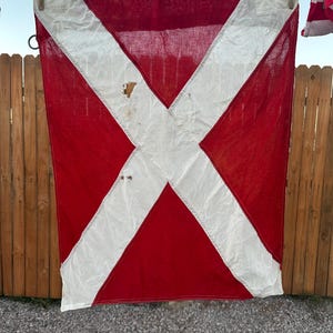 May include: A rectangular nautical signal flag featuring a large white diagonal cross on a solid red background. The fabric shows visible signs of wear, including small holes and light staining, with stitched seams along the edges.