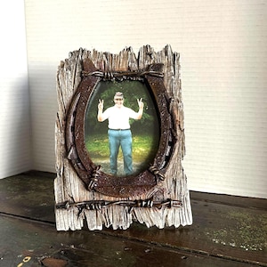 May include: A rustic picture frame with a horseshoe and barbed wire design. The frame surrounds a photo of a person. The frame is made of wood and has a weathered appearance, with a brown and gray color scheme.