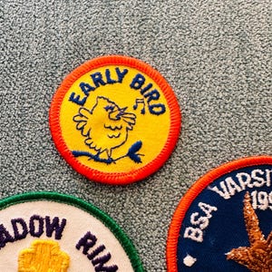 Op de afbeelding: Close-up van drie geborduurde patches. De bovenste patch heeft een gele kern met een blauwe vogel en de woorden "EARLY BIRD" in blauw. De tweede patch heeft een witte kern met de woorden "MEADOW RIM" en een gele bloem. De derde patch heeft een blauwe kern met de woorden "BSA VARSITY".