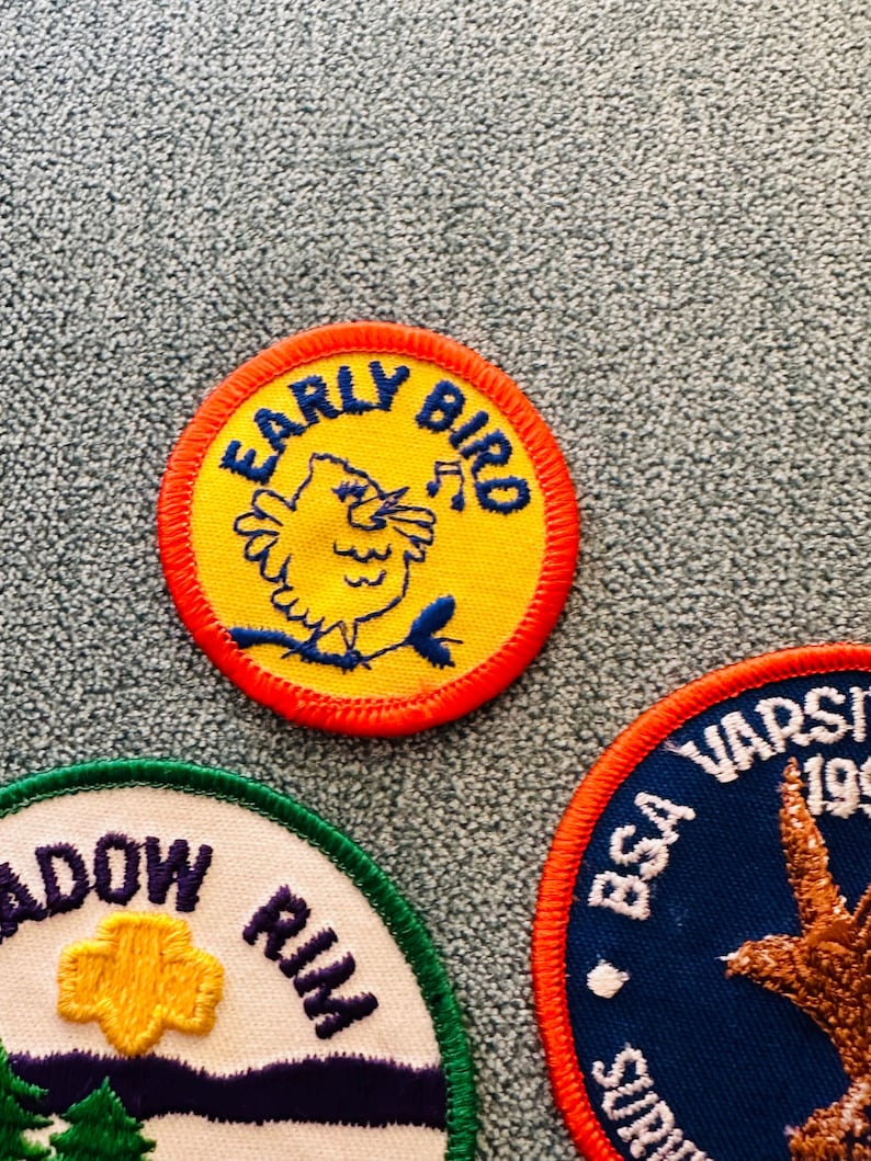 Op de afbeelding: Close-up van drie geborduurde patches. De bovenste patch heeft een gele kern met een blauwe vogel en de woorden "EARLY BIRD" in blauw. De tweede patch heeft een witte kern met de woorden "MEADOW RIM" en een gele bloem. De derde patch heeft een blauwe kern met de woorden "BSA VARSITY".