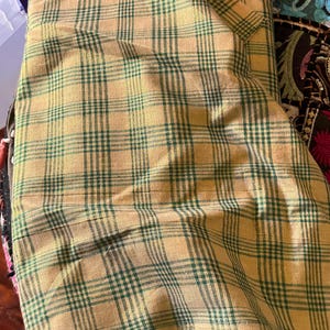 May include: A folded fabric with a plaid pattern in shades of tan and green. The fabric appears to be cotton or a similar material, with a classic check design. The fabric is folded over itself, showing the pattern in multiple layers.