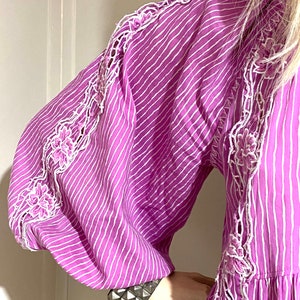 Puede incluir: Un vestido morado vibrante con rayas verticales blancas y un intrincado bordado floral blanco a lo largo de las mangas. El vestido presenta mangas voluminosas y un dise&ntilde;o escalonado. Se ve una pulsera plateada con tachuelas en la mu&ntilde;eca.