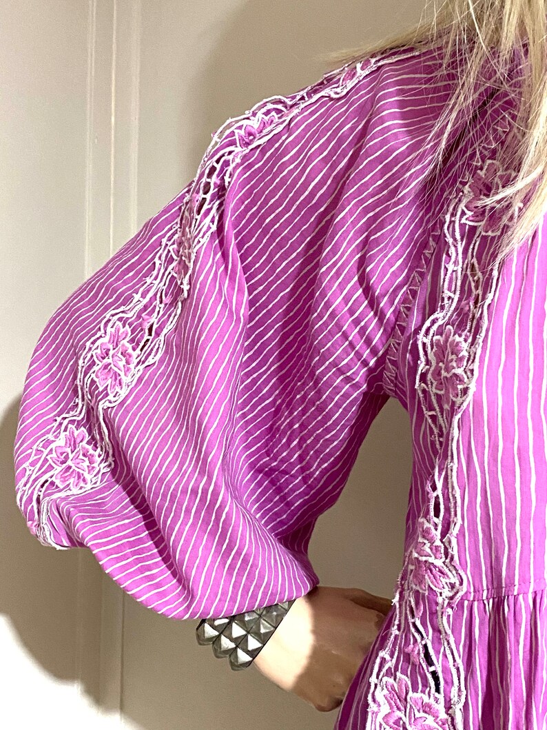 Puede incluir: Un vestido morado vibrante con rayas verticales blancas y un intrincado bordado floral blanco a lo largo de las mangas. El vestido presenta mangas voluminosas y un dise&ntilde;o escalonado. Se ve una pulsera plateada con tachuelas en la mu&ntilde;eca.