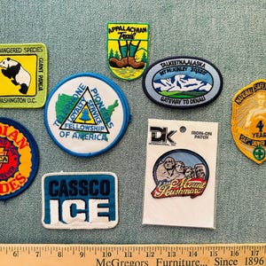 May include: A collection of eight embroidered patches featuring various designs including a panda, Appalachian Trail, Telephone Pioneers, Mount Rushmore, and YMCA Indian Guides. A wooden ruler is visible at the bottom for scale reference.