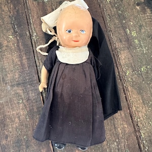 May include: A doll with a pale, smooth head, blue eyes, and a red mouth. It wears a black dress with short sleeves, a white collar, and a white headpiece with a long black veil attached. The doll has light-colored arms and black shoes.