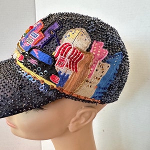 May include: A black baseball cap covered in dark sequins featuring an embroidered city skyline scene. The design includes colorful buildings, a blue bridge, and a red and white striped structure, all accented with intricate beadwork and stitching.