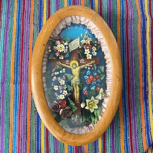 May include: An oval-shaped framed religious artwork featuring a depiction of Jesus on the cross, surrounded by colorful flowers. The frame is made of light-colored wood. The artwork includes the text "INRI" on a banner.