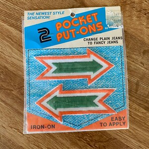 May include: A package of two iron-on pocket put-ons. The patches are arrow-shaped with orange and white borders and green centers. The package has text that reads "Pocket Put-Ons" and "Change Plain Jeans to Fancy Jeans."