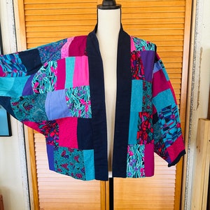 May include: A patchwork jacket with a navy blue collar and trim. The jacket features a variety of colorful fabric squares in shades of pink, teal, purple, and floral patterns. The jacket is displayed on a mannequin.
