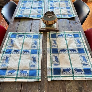 May include: Four rectangular placemats featuring a blue and green plaid pattern with repeating dark blue moose silhouettes and evergreen tree motifs. The word ALASKA is printed vertically along the side borders of each fabric placemat.