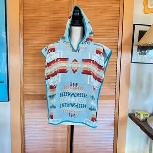 May include: A light blue hooded poncho with a Southwestern geometric pattern in red, orange, and white. The poncho has a V-neck and turquoise trim. The garment is displayed on a mannequin.
