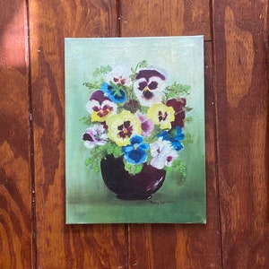 May include: An oil painting of a bouquet of pansies in a dark vase. The flowers are in shades of purple, yellow, blue, and white, set against a light green background. The painting is signed by the artist, Ruby Williamson.