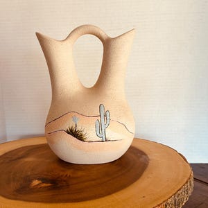 May include: A tan ceramic wedding vase with a textured matte finish, featuring a double-spout design and a central arched handle. The front is hand-painted with a desert landscape scene including a light blue saguaro cactus and rolling hills.