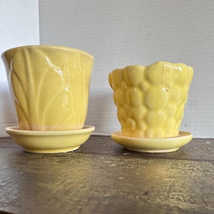 May include: Two yellow ceramic flower pots with matching saucers. One pot has a textured leaf design, the other has a bubble-like pattern. Both are vintage and sit on a wooden surface.