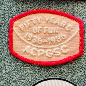 Op de afbeelding: Geborduurde patch met de tekst "FIFTY YEARS OF FUN 1936-1986 ACPGSC" in witte draad op een perzikkleurige achtergrond. De patch heeft een felroze rand, waarschijnlijk een vintage verzamelobject of souvenir.