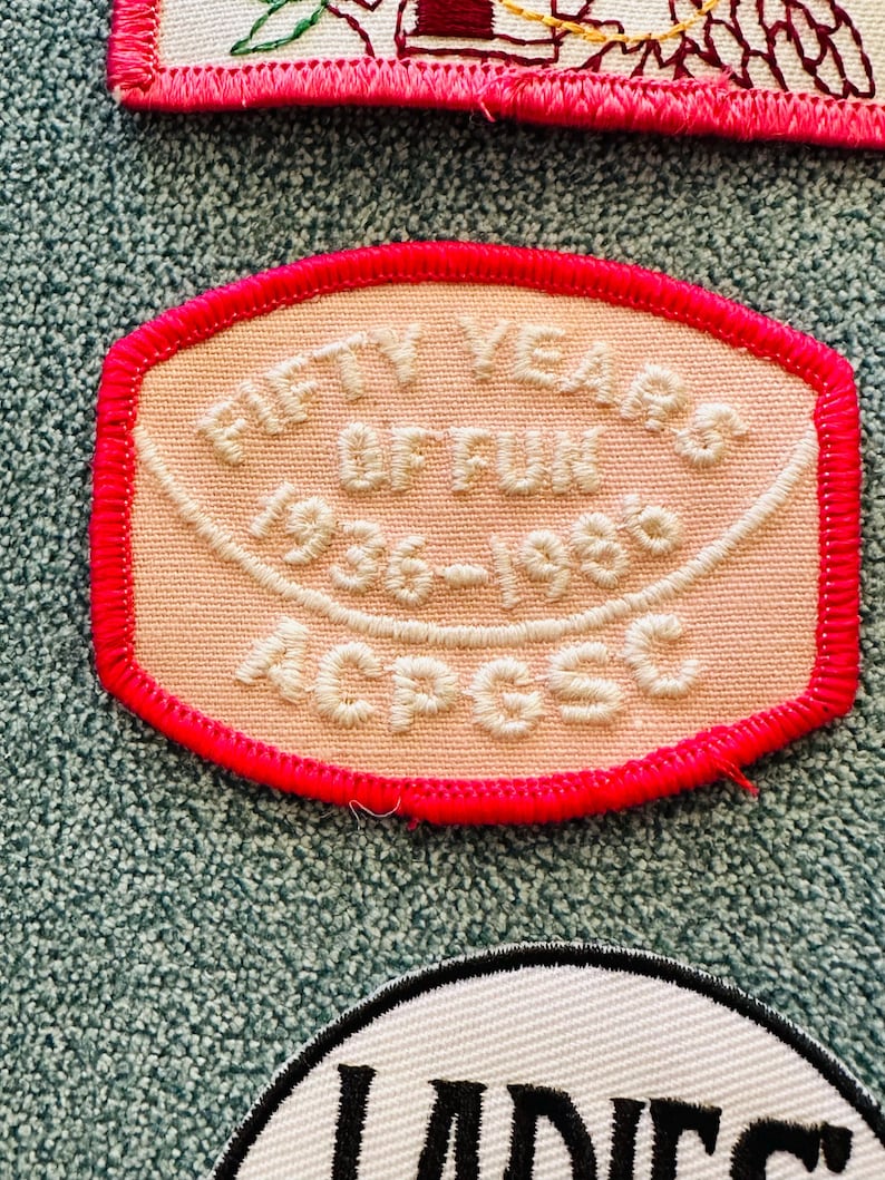 Op de afbeelding: Geborduurde patch met de tekst "FIFTY YEARS OF FUN 1936-1986 ACPGSC" in witte draad op een perzikkleurige achtergrond. De patch heeft een felroze rand, waarschijnlijk een vintage verzamelobject of souvenir.