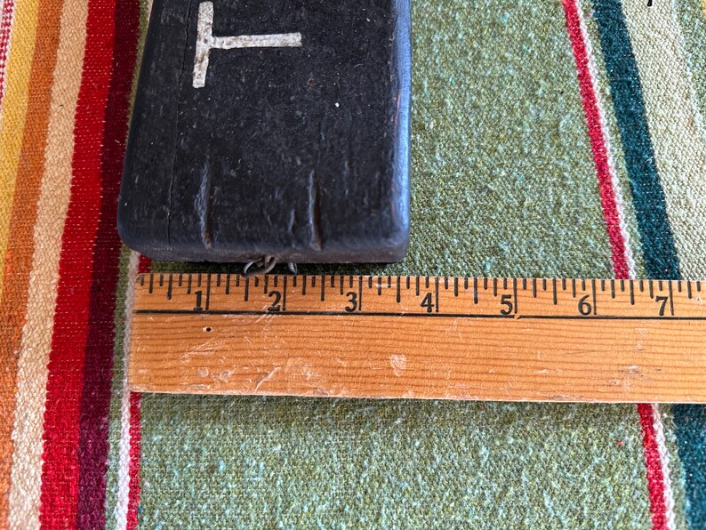 Op de afbeelding: Een donker, rechthoekig object met een witte "T"-vorm aan de bovenkant, naast een houten liniaal. De liniaal toont metingen in centimeters. Het object lijkt gemaakt van een donker, mogelijk houten materiaal.